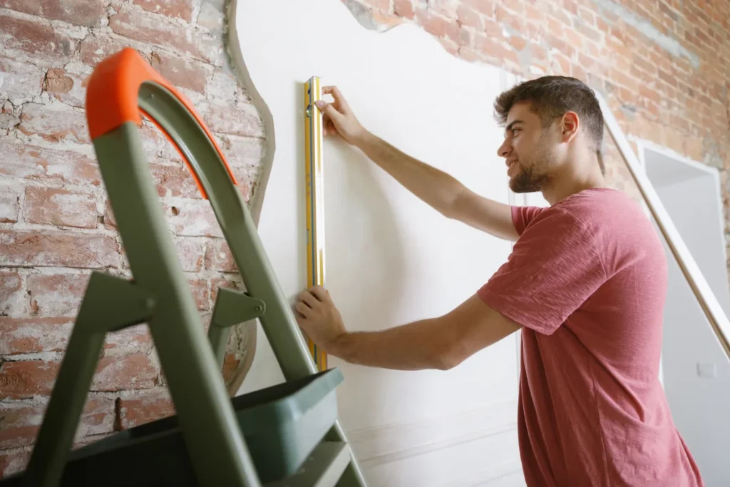 Hombre midiendo una pared con un nivel mientras realiza trabajos de remodelación en un ambiente con ladrillos expuestos y una escalera de mano.