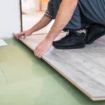 Hombre instalando un piso laminado de color claro en una habitación, asegurando las piezas con las manos sobre un suelo verde.